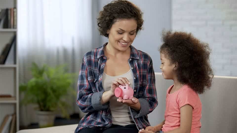 Mother and daughter sitting on couch placing coins in piggy bank