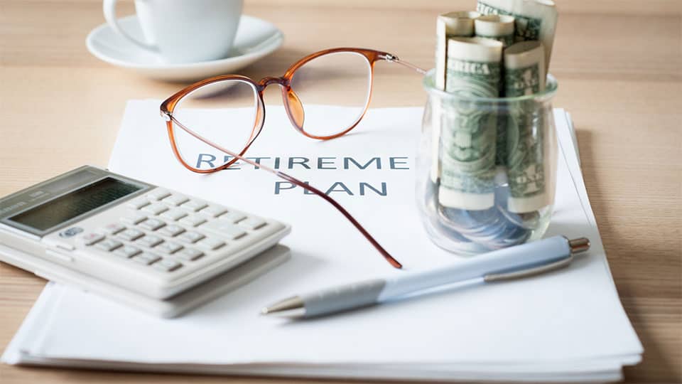 Un documento sobre la mesa con la etiqueta &quot;Plan de jubilación&quot; debajo de unas gafas, una calculadora, un bolígrafo y un pequeño tarro de cristal con billetes de dólar y monedas en su interior.