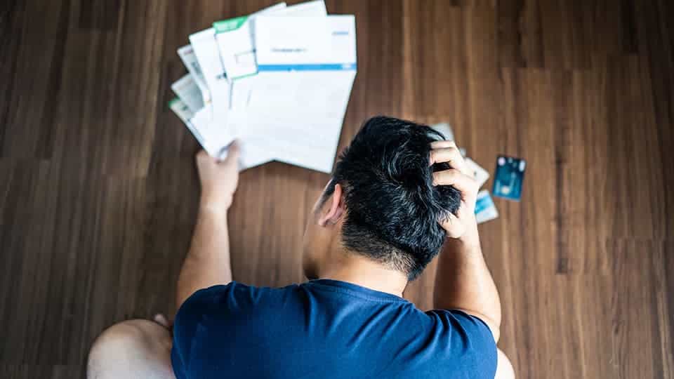 Frustrated person holding bills in one hand with credit card thrown about on wooden floor