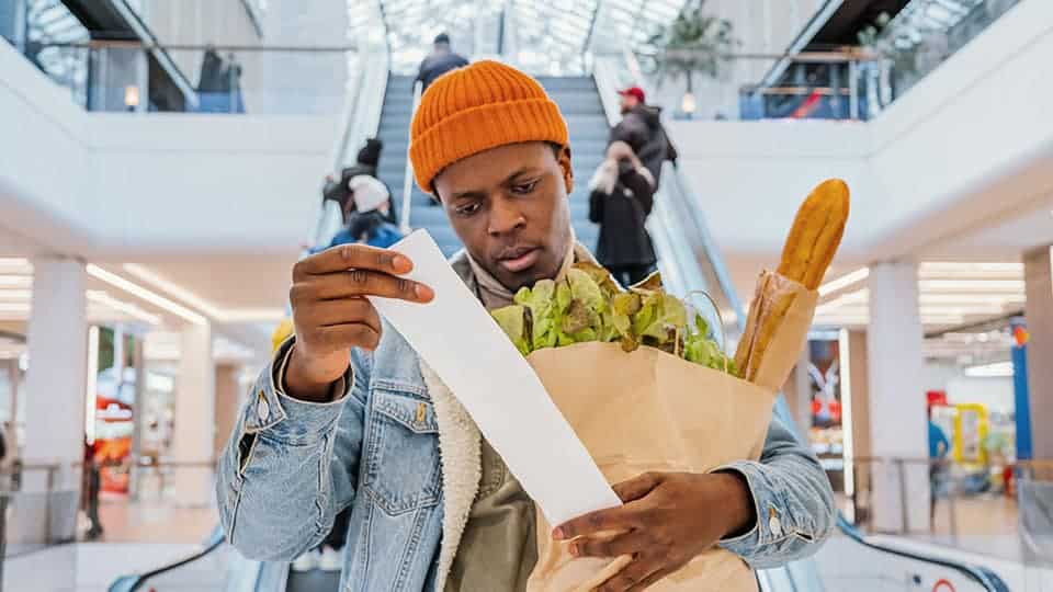 Man with large bag of groceries under arm looking at long receipt