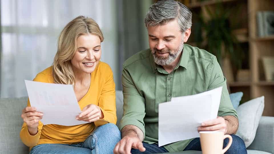 Couple sitting next to eachother on couch holding documents in both hands and looking at screen on coffee table