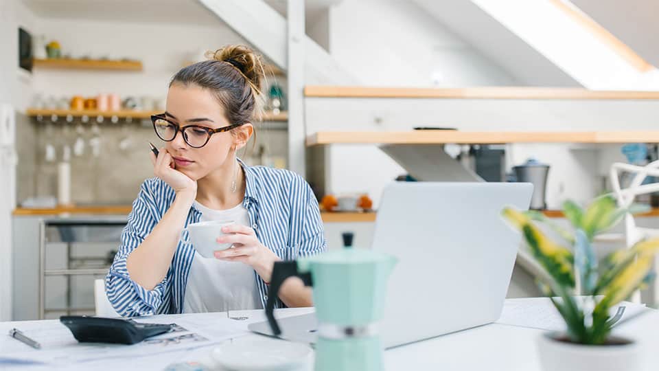 Mujer sentada en la mesa de la cocina con una taza de café en la mano y mirando los papeles con el ordenador portátil y la planta que descansa sobre la superficie a su lado