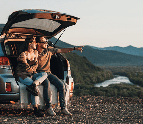 Man and woman sitting in the back of car looking out over mountainscape at sunset