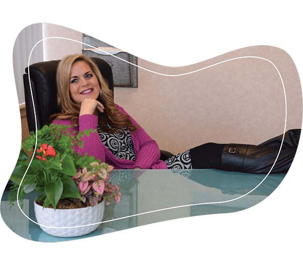 Jennifer Denoo sitting comfortably in office with feet propped up on desk smiling for camera with plant sitting on table in front of her