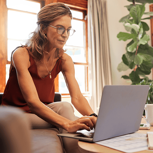 Lady sitting on couch in living room working on laptop with bright light and plants behind her