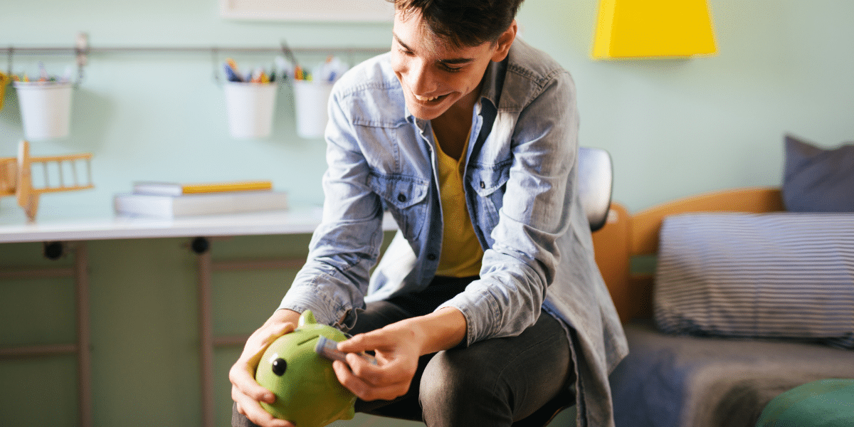 Young teenage boy in a denim jacket putting a bill into a green piggy bank while sitting on his bed
