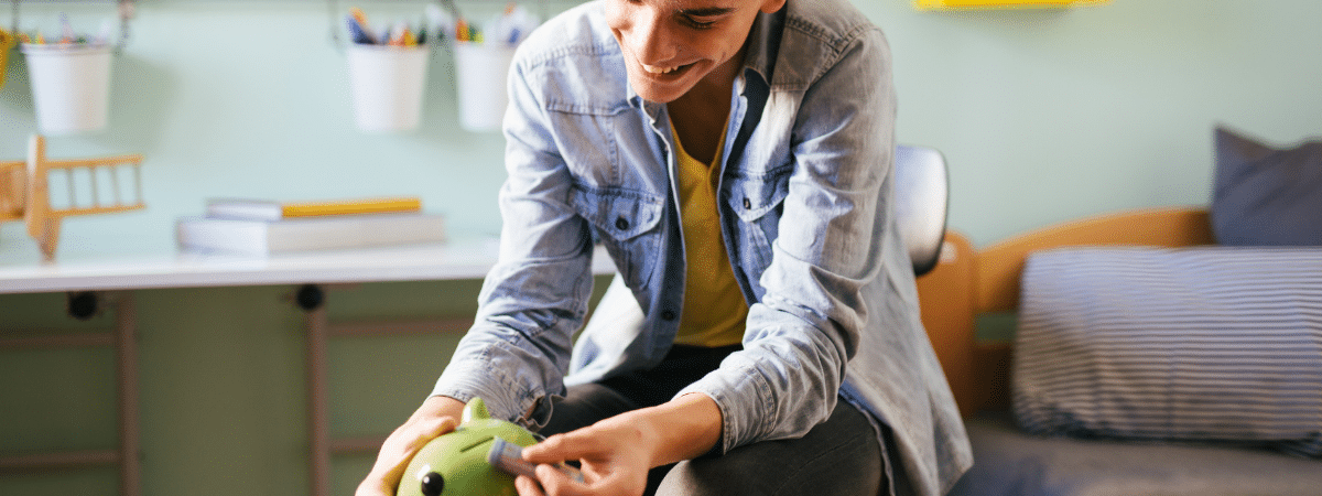 Young teenage boy in a denim jacket putting a bill into a green piggy bank while sitting on his bed